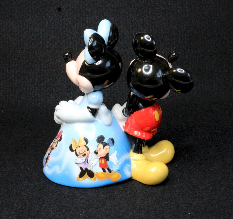 May include: A ceramic figurine of Mickey Mouse and Minnie Mouse. Mickey is standing behind Minnie, who is wearing a blue dress with a white and blue pattern. The figurine is painted in bright colors and features a whimsical design.