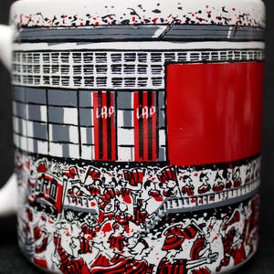 May include: White ceramic mug with a stadium design. The mug features a red, black, and white illustration of a crowd of people, flags, and stadium architecture. The word "LAP" is visible on the mug.