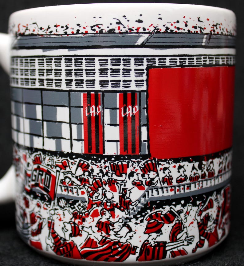 May include: White ceramic mug with a stadium design. The mug features a red, black, and white illustration of a crowd of people, flags, and stadium architecture. The word "LAP" is visible on the mug.
