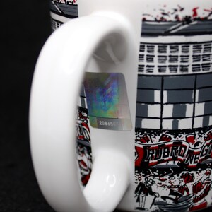 May include: White ceramic mug with a stadium design in gray, black, and red. The mug features a curved handle and a holographic sticker with the number 2086505. The design includes the text "RUBRO NE".