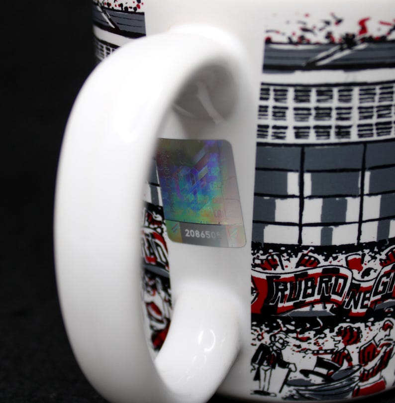 May include: White ceramic mug with a stadium design in gray, black, and red. The mug features a curved handle and a holographic sticker with the number 2086505. The design includes the text "RUBRO NE".