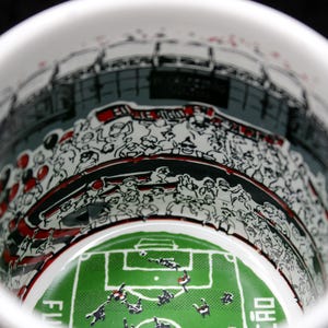 May include: White ceramic mug with a detailed illustration of a soccer stadium interior. The design includes a green field, players, and a crowd of spectators in black, white, and red. The mug's interior is visible.