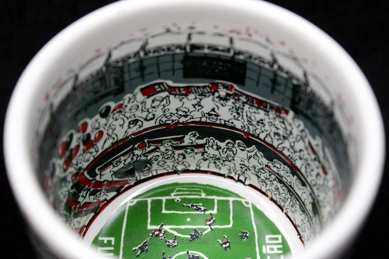May include: White ceramic mug with a detailed illustration of a soccer stadium interior. The design includes a green field, players, and a crowd of spectators in black, white, and red. The mug's interior is visible.
