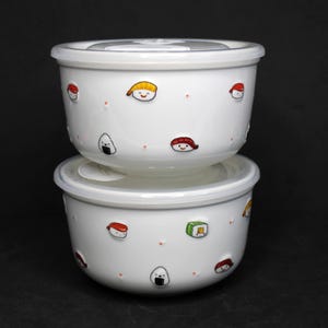 May include: Two white ceramic bowls with lids stacked on top of each other. The bowls are decorated with cartoon sushi characters and small orange dots. The lids are light gray. The background is black.