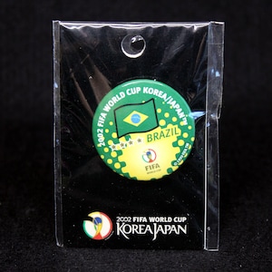 May include: A pin featuring the Brazilian flag and the text "BRAZIL" on a green and yellow background. The pin commemorates the 2002 FIFA World Cup Korea/Japan. The pin is in a clear plastic package.
