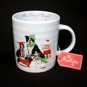 May include: A white ceramic mug with a winter scene featuring dogs wearing hats and scarves. The mug has the text "All is Furry & Bright!" and a red tag that says "The Mistletoe Co."