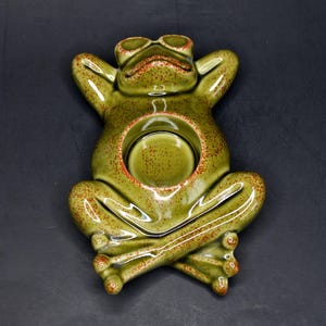 May include: A ceramic frog-shaped candle holder in a relaxed pose. The frog is glazed in shades of green with reddish-brown speckles and accents. The candle holder has a circular opening in its belly to hold a candle.