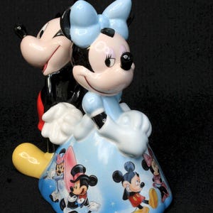 May include: A ceramic figurine of Mickey Mouse and Minnie Mouse. Mickey is wearing his signature red shorts and black shoes. Minnie is wearing a blue dress with a white bow. The figurine is decorated with a blue background and images of Mickey and Minnie in different poses.