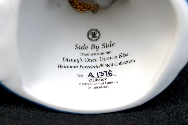 May include: White porcelain bell with a gold chain, featuring the text "Side By Side", "Third issue in the Disney's Once Upon a Kiss Heirloom Porcelain Bell Collection", "No. A 1376", "&copy;Disney", "&copy;2005 Bradford Editions", and "15-16839-003".