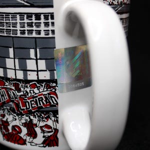 May include: White ceramic mug with a wraparound design featuring a stadium scene in gray and black, with red accents and text. The mug has a white handle and a holographic sticker with the number 2086505.