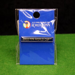 May include: A rectangular metal pin featuring the text 2002 FIFA WORLD CUP in gold lettering. The pin is attached to a blue display card with the 2002 FIFA WORLD CUP KOREA JAPAN logo and text, all enclosed in a clear plastic protective sleeve.