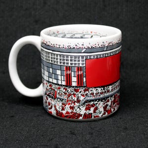 May include: White ceramic coffee mug with a wraparound design depicting a stadium scene with red, gray, and white elements. The mug features a large red section and a handle on the side.