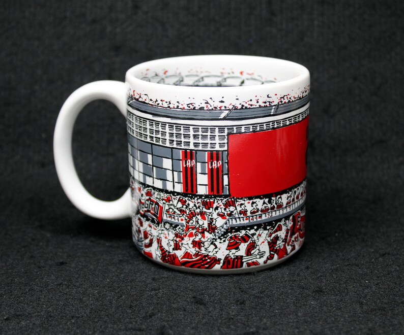 May include: White ceramic coffee mug with a wraparound design depicting a stadium scene with red, gray, and white elements. The mug features a large red section and a handle on the side.
