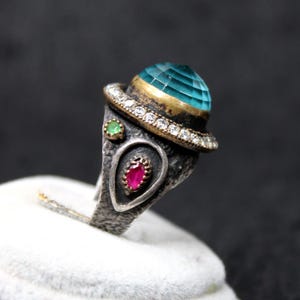 May include: A textured dark metal ring featuring a large, faceted, round teal stone set in a gold-toned bezel, surrounded by a halo of small clear stones. The band includes a small round green stone and a pear-shaped red stone in a silver setting.
