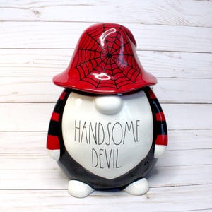 May include: A black and red ceramic gnome figurine with a spiderweb hat. The gnome is wearing a black and red striped shirt and has the words "Handsome Devil" written on its chest.
