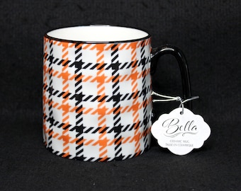 May include: A white ceramic mug featuring an orange and black houndstooth plaid pattern. The mug has a black handle and a scalloped white paper tag attached with a silver string. The tag displays the text Bella, Ceramic Mug, and Tasse en Céramique.
