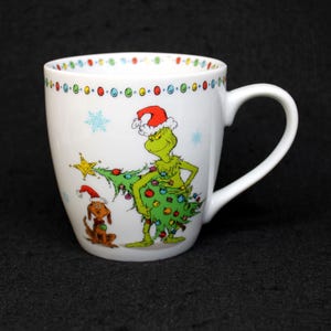 May include: White ceramic mug featuring a Christmas-themed illustration of the Grinch holding a decorated Christmas tree, with a dog wearing a Santa hat. The mug has a white handle and a rim decorated with colorful dots.
