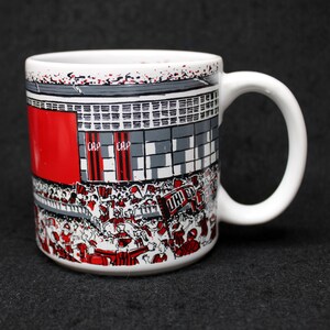 May include: White ceramic mug featuring a stadium scene with red, gray, and white details. The design includes a crowd of people, banners, and the text "CAP". The mug has a white handle.