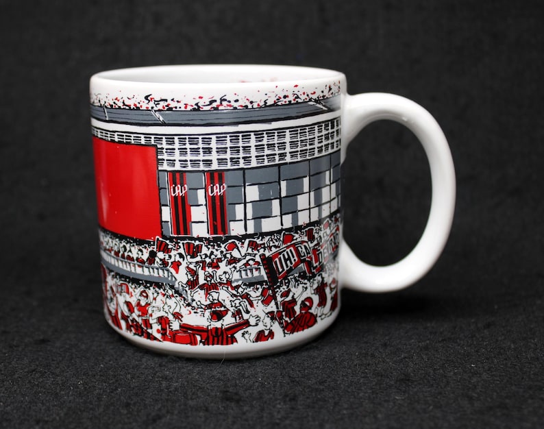 May include: White ceramic mug featuring a stadium scene with red, gray, and white details. The design includes a crowd of people, banners, and the text "CAP". The mug has a white handle.