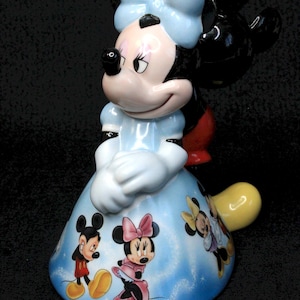 May include: A ceramic figurine of Minnie Mouse wearing a blue dress with a white bow. The dress is decorated with images of Mickey Mouse, Minnie Mouse, and Donald Duck.