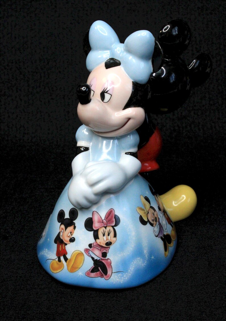 May include: A ceramic figurine of Minnie Mouse wearing a blue dress with a white bow. The dress is decorated with images of Mickey Mouse, Minnie Mouse, and Donald Duck.