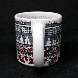 May include: White ceramic mug with a wraparound design featuring a stadium scene in gray, black, and red. The design includes text and illustrations of people. The mug has a white handle and a black background.