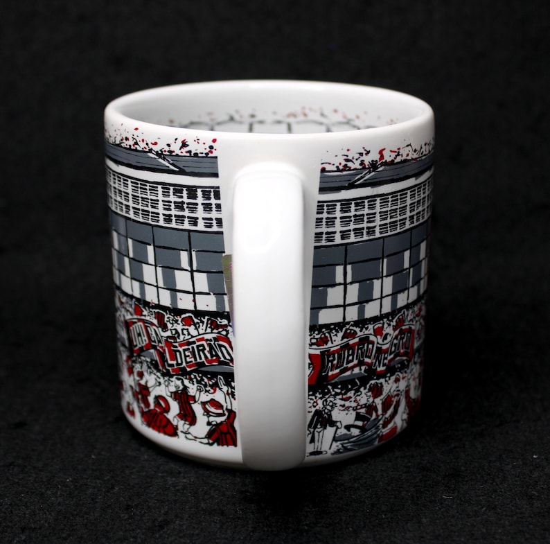 May include: White ceramic mug with a wraparound design featuring a stadium scene in gray, black, and red. The design includes text and illustrations of people. The mug has a white handle and a black background.