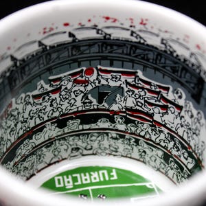 May include: White ceramic mug with a stadium illustration. The interior features a crowd of people with flags, a stadium structure, and a green field with the word "FURAC&Atilde;O". Red accents are visible.
