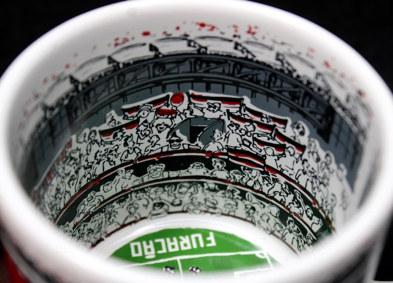 May include: White ceramic mug with a stadium illustration. The interior features a crowd of people with flags, a stadium structure, and a green field with the word "FURAC&Atilde;O". Red accents are visible.