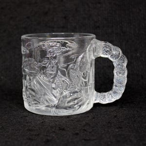 May include: Clear glass mug with a detailed embossed design. The mug features a portrait and other imagery. The handle is made of a series of coin-like shapes. The mug is transparent and sits on a dark surface.