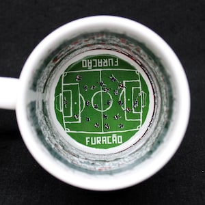 May include: White ceramic mug with a soccer field design inside. The green field features players and the word "FURAC&Atilde;O" in white. The mug has a white handle and a black background.