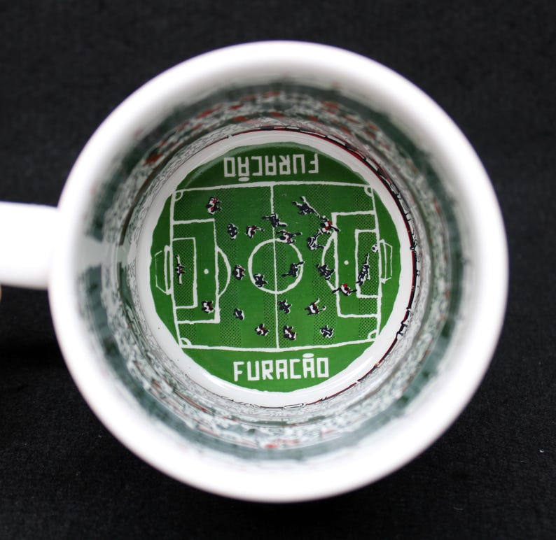 May include: White ceramic mug with a soccer field design inside. The green field features players and the word "FURAC&Atilde;O" in white. The mug has a white handle and a black background.