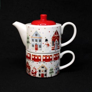 May include: A white ceramic Christmas-themed tea set with a red lid. The teapot sits atop a matching teacup. The set is decorated with images of houses, a snowman, and a train. The set is perfect for the holiday season.
