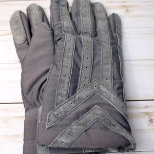 May include: A pair of grey gloves with suede detailing. The gloves feature a quilted design with suede stripes and a chevron pattern on the back. The suede is a lighter shade of grey than the main glove material.