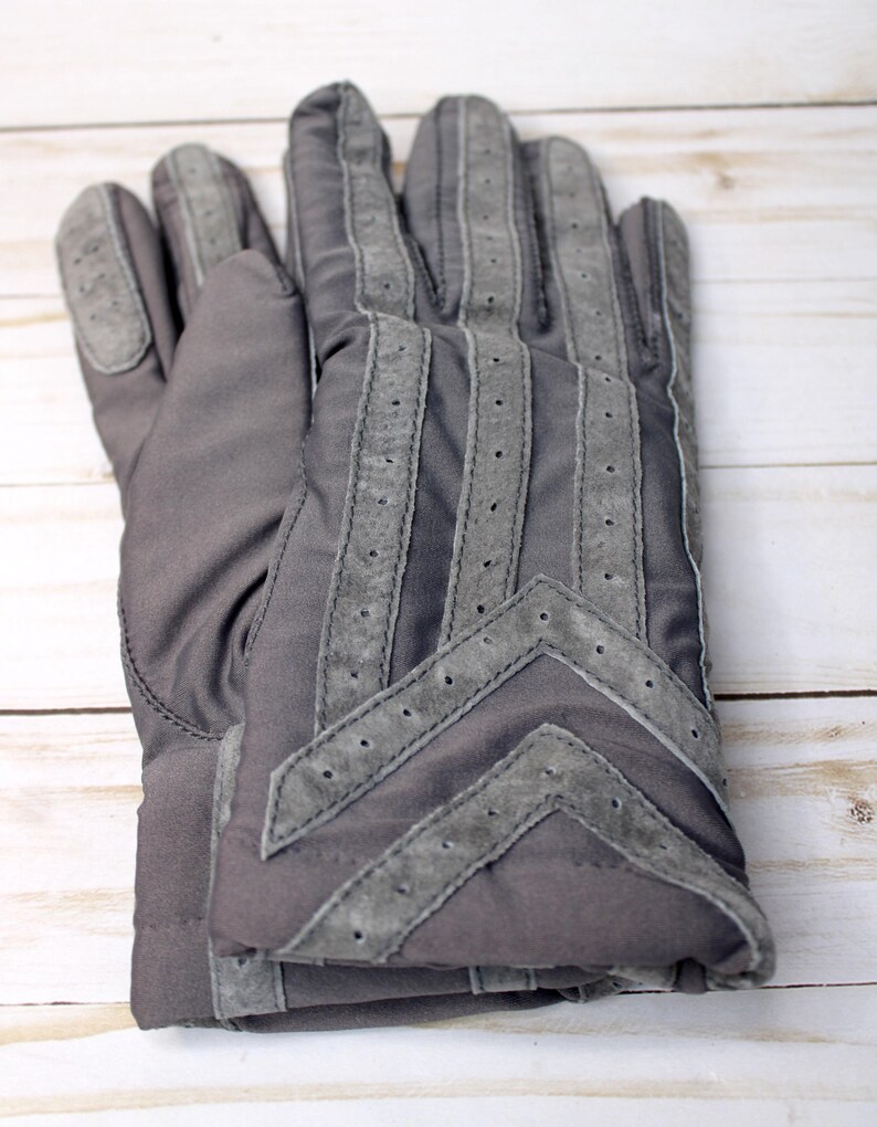 May include: A pair of grey gloves with suede detailing. The gloves feature a quilted design with suede stripes and a chevron pattern on the back. The suede is a lighter shade of grey than the main glove material.