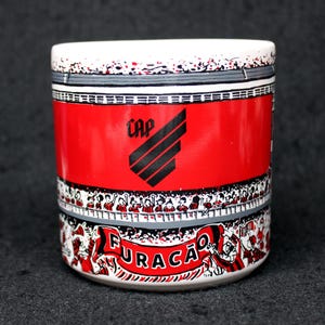 May include: A cylindrical ceramic mug with a white rim and base, featuring a red central band with the word "CAP" and a black logo. The design includes a crowd of people and the word "FURAC&Atilde;O" in a banner.