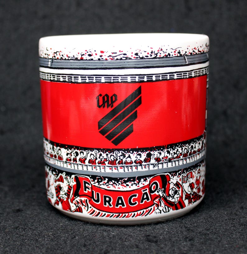 May include: A cylindrical ceramic mug with a white rim and base, featuring a red central band with the word "CAP" and a black logo. The design includes a crowd of people and the word "FURAC&Atilde;O" in a banner.