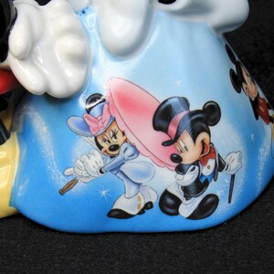 May include: A ceramic figurine of Mickey Mouse and Minnie Mouse. Mickey is wearing red shorts and yellow shoes. Minnie is wearing a blue dress with white polka dots and a red bow. The figurine is decorated with a blue background and images of Mickey and Minnie in different poses.