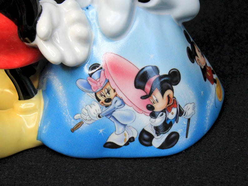 May include: A ceramic figurine of Mickey Mouse and Minnie Mouse. Mickey is wearing red shorts and yellow shoes. Minnie is wearing a blue dress with white polka dots and a red bow. The figurine is decorated with a blue background and images of Mickey and Minnie in different poses.