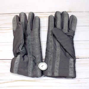 May include: A pair of dark grey gloves with suede-like accents. The gloves feature vertical grey stripes with small perforations and a cuff detail. The gloves are displayed on a white wooden surface.