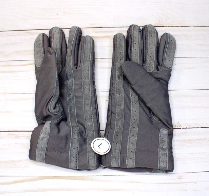 May include: A pair of dark grey gloves with suede-like accents. The gloves feature vertical grey stripes with small perforations and a cuff detail. The gloves are displayed on a white wooden surface.