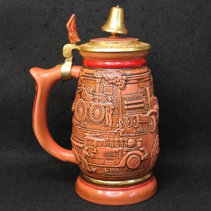 May include: A ceramic beer stein with a reddish-brown finish featuring embossed historical fire truck imagery and the years 1822, 1917, and 1939. It includes a metallic gold-colored lid topped with a small bell and a matching handle with a thumb lift.