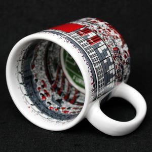 May include: White ceramic mug with a wraparound design featuring a cityscape in shades of gray, red, and white. The mug has a curved handle and a glossy finish. The interior of the mug is visible, with a green and white design.