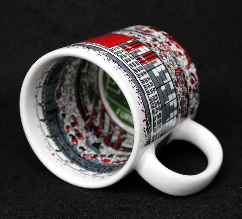 May include: White ceramic mug with a wraparound design featuring a cityscape in shades of gray, red, and white. The mug has a curved handle and a glossy finish. The interior of the mug is visible, with a green and white design.