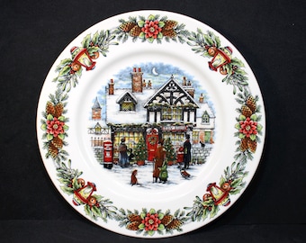 May include: A white ceramic Christmas plate with a festive winter scene. The plate features a snow-covered building with a red door and a sign that reads "Toy Shop". The building is surrounded by snow-covered trees and a red mailbox. The plate is decorated with a green holly and red berry border.