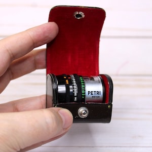 May include: A Petri Meter camera lens in a brown leather case with a red velvet interior. The lens has black and silver details with measurement markings. The case is held open by a hand.
