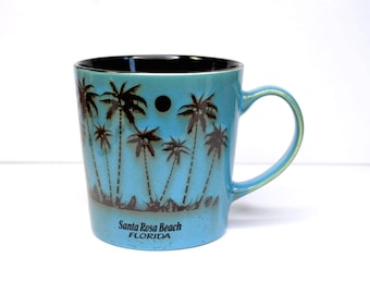 May include: A blue ceramic mug with a black rim and handle. The mug features a black silhouette of palm trees against a blue background. The text "Santa Rosa Beach FLORIDA" is printed on the bottom of the mug.