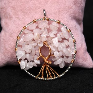 May include: A handcrafted pendant featuring a tree of life design. The pendant has a silver-colored outer ring and copper-colored wire forming the tree's trunk and branches. The tree's leaves are made of small, light pink rose quartz stones.