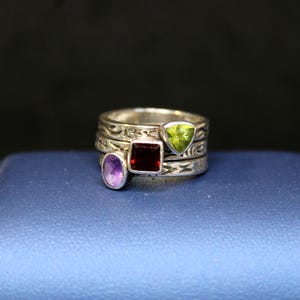 May include: A stack of silver rings with decorative etched bands. Each ring features a different gemstone: an oval purple stone, a square red stone, and a triangular green stone. The rings are displayed on a blue surface.