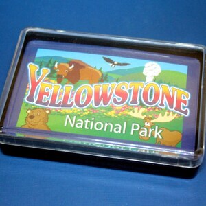 May include: A clear plastic frame with a colorful image of Yellowstone National Park. The image features a bison, a bear, a moose, and a bald eagle. The text "Yellowstone National Park" is printed in yellow, orange, and red letters.